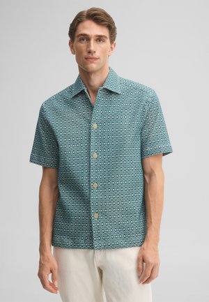 Homme portant une chemise verte à motifs, à manches courtes et boutonnée, avec un pantalon de couleur claire, debout devant un fond uni.