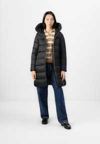 Zwarte pufferjas met capuchon, voorzien van een rits aan de voorkant en een gewatteerde textuur. Draag met een beige gestreepte trui en een relaxed paar blauwe jeans.