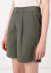 Shorts vert olive en tissu léger, dotés d'une taille avec plis, de poches latérales et d'une coupe droite. Les finitions à l'ourlet sont soignées.