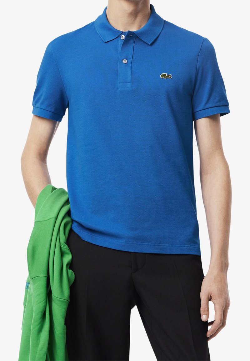Homme portant un polo Lacoste bleu et un pantalon noir, tenant une veste verte dans la main gauche sur fond blanc.