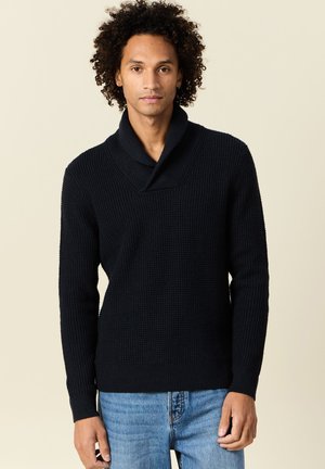 Jersey de punto en azul marino oscuro con cuello de chal, superficie texturizada, mangas largas y puños acanalados, combinado con jeans de color azul claro.