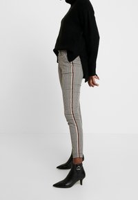 Pantalons fuselés à motif pied-de-poule avec une bande latérale rouge et blanche, associés à des bottines noires à talon pointu et un pull noir ample.