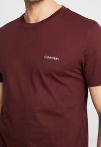 Bordeaux katoenen t-shirt met een ronde hals, korte mouwen en een klein wit "Calvin Klein" logo op de linkerborst. Soepele textuur.