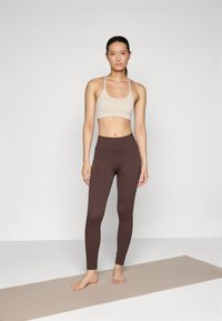 Conjunto de yoga composto por um sutiã desportivo bege claro e leggings de cintura alta castanhas escuras, ambos feitos de um tecido elástico, sobre um tapete bege.