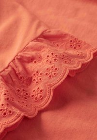 Vêtement en coton corail avec un ourlet à volants et des détails de broderie florale. Le tissu a une texture douce avec une finition lisse.