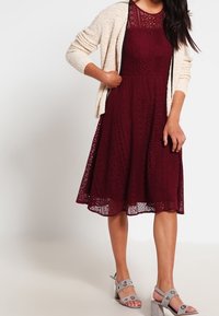 Burgunderfärgad spetsklänning med åtsittande midja och utsvängd kjol, som matchas med en ljuskrämfärgad cardigan och grå sandaler med silverdetaljer.