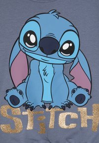Blå grafisk hettegenser med Stitch, en tegneseriefigur, med store uttrykksfulle øyne, rosa detaljer på ørene og gull-glitrende tekst som sier "Stitch."