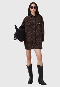 Abito camicia marrone a stampa leopardata con bottoni e tasche frontali, abbinato a una borsa nera e stivali da cowboy neri al ginocchio.