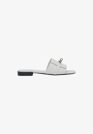 White flat sandal with a minimalist design, featuring a wide strap and a circular accent. Smooth leather material with a black sole.
