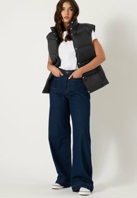 Gilet rembourré noir sans manches, porté sur un t-shirt blanc. Associé à un jean large bleu foncé et des baskets blanches.