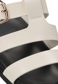 Primer plano de una sandalia de cuero beige con dos tiras y una hebilla dorada sobre una suela negra.