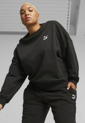 Sweatshirt nero oversize con texture a coste e logo Puma bianco sul petto, abbinato a pantaloni jogger grigi scuri con tasche laterali.
