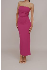 Abito maxi aderente senza spalline in tessuto rosa brillante, caratterizzato da una texture liscia e un design che arriva fino al pavimento. Abbinato a sandali dorati con cinturini.