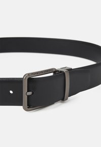 Ceinture noire avec une finition texturée, dotée d'une boucle en métal avec un logo gravé. La boucle a une teinte argentée douce et un design classique.