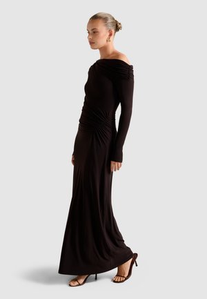 Woman in a long black off-shoulder dress with ruched details, wearing brown high-heeled sandals and gold earrings, standing in profile.