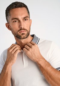 Vit polo-shirt med en texturerad grå krage och mörkblå detaljer. Modellen justerar kragen och visar den knäppta halsen.