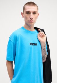 Tyrkysové tričko s krátkym rukávom z bavlny, so čiernym logom na hrudi. Čierna bunda je prehodená cez rameno modelky.
