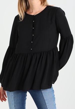 Femme portant un chemisier noir à manches longues avec des détails de boutons et une taille froncée, associé à un jean bleu sur un fond uni.