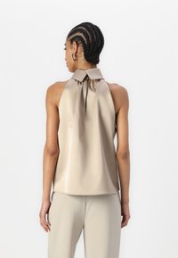 Top senza maniche in raso beige chiaro con colletto alto e dettaglio annodato. Texture liscia, design fluido, vista posteriore, abbinato a pantaloni chiari.