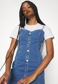 Denim dress with button front, fitted design, and adjustable straps, layered over a white T-shirt. Textured blue fabric.