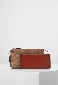 Brązowa torebka kopertówka z wzorem na płótnie, z czerwoną skórzaną kieszonką akcentującą. Posiada złote okucia i zamek błyskawiczny, oraz odpinany pasek na nadgarstek.