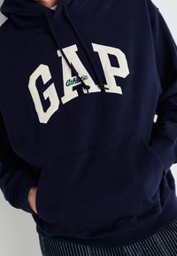 Felpa con cappuccio blu navy con la scritta bianca "GAP" e dettagli verdi. Presenta una tasca frontale e cappuccio con cordini, realizzata in tessuto morbido.