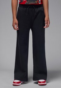 Pantalon de survêtement noir à jambes larges avec une taille élastique et un cordon de serrage. Le tissu est doux et l'ourlet est fini par un subtil revers.