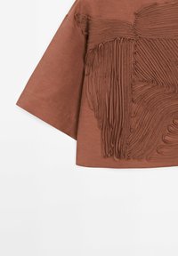 Haut marron à manches courtes en tissu texturé, présentant un design plissé détaillé sur le devant et des manches lisses.