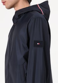 Marineblauwe hoodiejas van lichtgewicht materiaal met een ritssluiting, zijzakken en een logopatch op de mouw met rode en witte accenten.