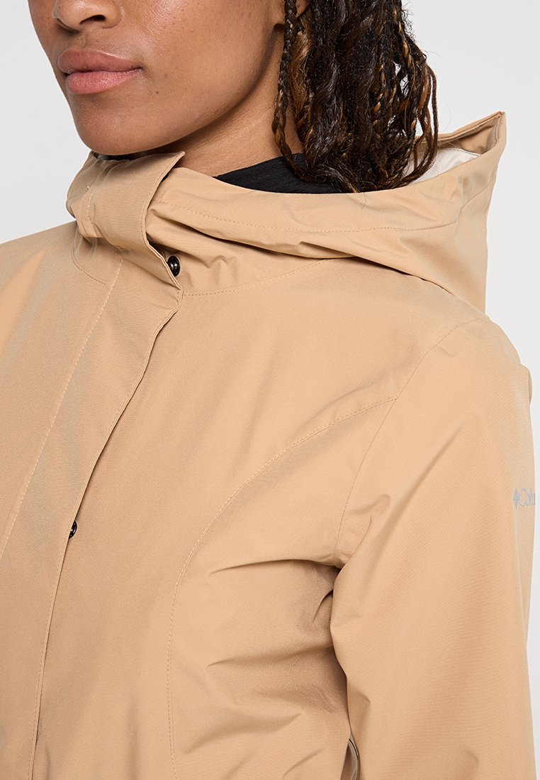Beżowa kurtka na zewnątrz z wysokim kołnierzem i regulowanym kapturem. Charakteryzuje się gładką, lekką teksturą oraz subtelnym logo z boku.
