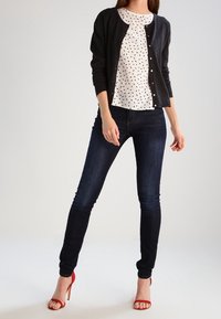 Cardigan nero, blusa bianca a fantasia, jeans scuri, sandali con tacco rosso. Presenta chiusura con bottoni e design aderente.