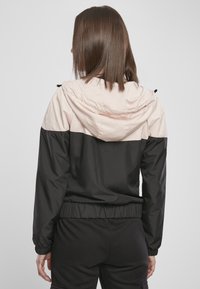 Giacca con cappuccio nera e rosa chiaro con polsini e vita elasticizzati, caratterizzata da una texture liscia e da blocchi di colore a contrasto.