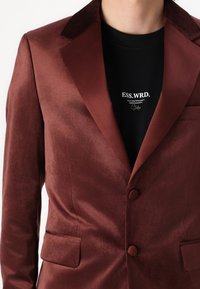 Blazer burdeos de tela suave, con solapas muestradas y dos bolsillos delanteros. Lleva puesto una camisa negra con texto blanco.