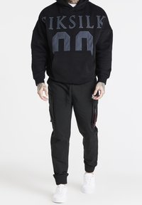 Felpa nera con 'SIKSILK 00' stampato sul davanti; abbinata a pantaloni jogger neri con tasche cargo e polsini elasticizzati. Sneakers bianche.