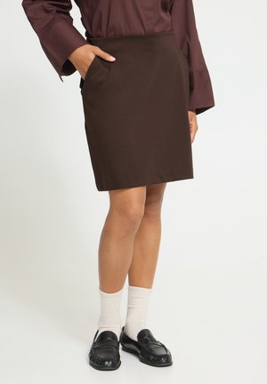 Femme portant une jupe trapèze marron foncé, un chemisier bordeaux à manches longues, des chaussettes blanches et des mocassins noirs, avec une main dans la poche de la jupe.