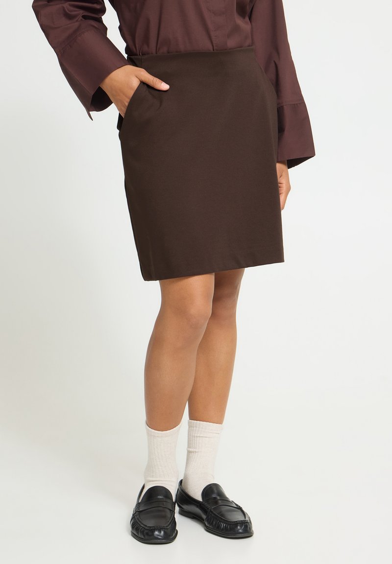 Femme portant une jupe trapèze marron foncé, un chemisier bordeaux à manches longues, des chaussettes blanches et des mocassins noirs, avec une main dans la poche de la jupe.