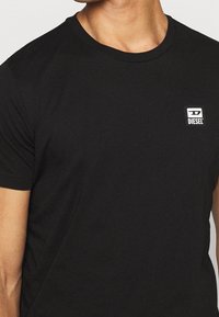 Mann trägt ein schlichtes schwarzes Diesel-T-Shirt mit einem kleinen weißen und schwarzen Diesel-Logo auf der linken Brust, gezeigt von Hals bis Taille.