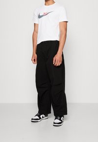 Bílý Nike tričko s šedým a červeným logem Swoosh, černé volné kalhoty a černobílé Nike tenisky, představující neformální atletický outfit.