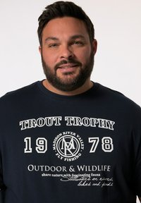 Námořnická modrá košile s dlouhým rukávem s výrazným bílým textem "TROUT TROPHY 1978" a grafikou se rybářskou tématikou. Volný styl, texturovaný materiál.