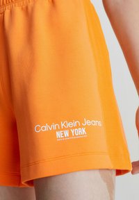 Oranssit puuvillashortsit, joissa on ribbitekstuuri ja valkoisella printillä teksti "Calvin Klein Jeans NEW YORK" alareunassa.