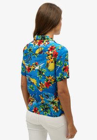 Camisa de manga corta hecha de una tela ligera que presenta un fondo azul vibrante con flores tropicales, palmeras y olas.