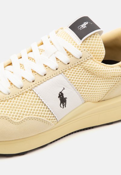 Baskets beige en mesh et suède avec lacets blancs, logo joueur de polo noir sur le panneau latéral blanc et la languette, et semelle beige clair.