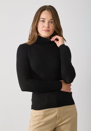 Black long-sleeve turtleneck top with a fitted design. Soft fabric and seamless cuffs. Worn with light-colored, casual pants.