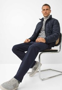 Mannen navy gewatteerde jas, coltrui met geometrisch patroon, navy broek en grijze sneakers, zittend op een chroomstoel.