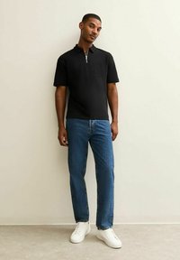 Zwarte polo t-shirt met korte mouwen, een ritskraag en een borstzak aan de linkerzijde, gecombineerd met blauwe denim jeans en witte sneakers op een neutrale achtergrond.