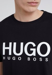 Zwart katoenen T-shirt met grote witte tekst "HUGO" en daaronder "HUGO BOSS". Ronde hals en korte mouwen. Soepele textuur.