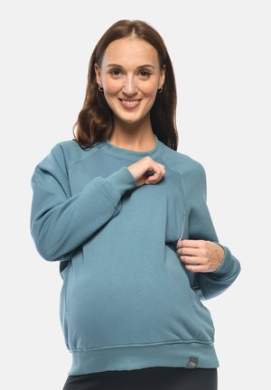 Lächelnde schwangere Frau trägt einen blauen Sweatshirt und öffnet einen seitlichen Reißverschluss, der für den Stillzugang konzipiert ist.