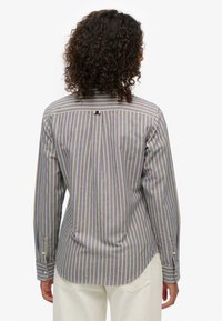 Person med krøllet hår iført en langærmet blå skjorte med vertikale beige striber, set bagfra, kombineret med hvide bukser.