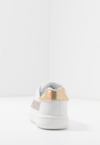 Zapatilla blanca con un acabado suave, que presenta un acento metálico dorado en el talón y un logo en un tono rosa suave en el costado.