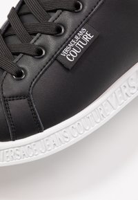 Černé syntetické tenisky s bílou gumovou podrážkou. Mezi vlastnosti patří šněrování, texturovaný svršek a logo na boku.